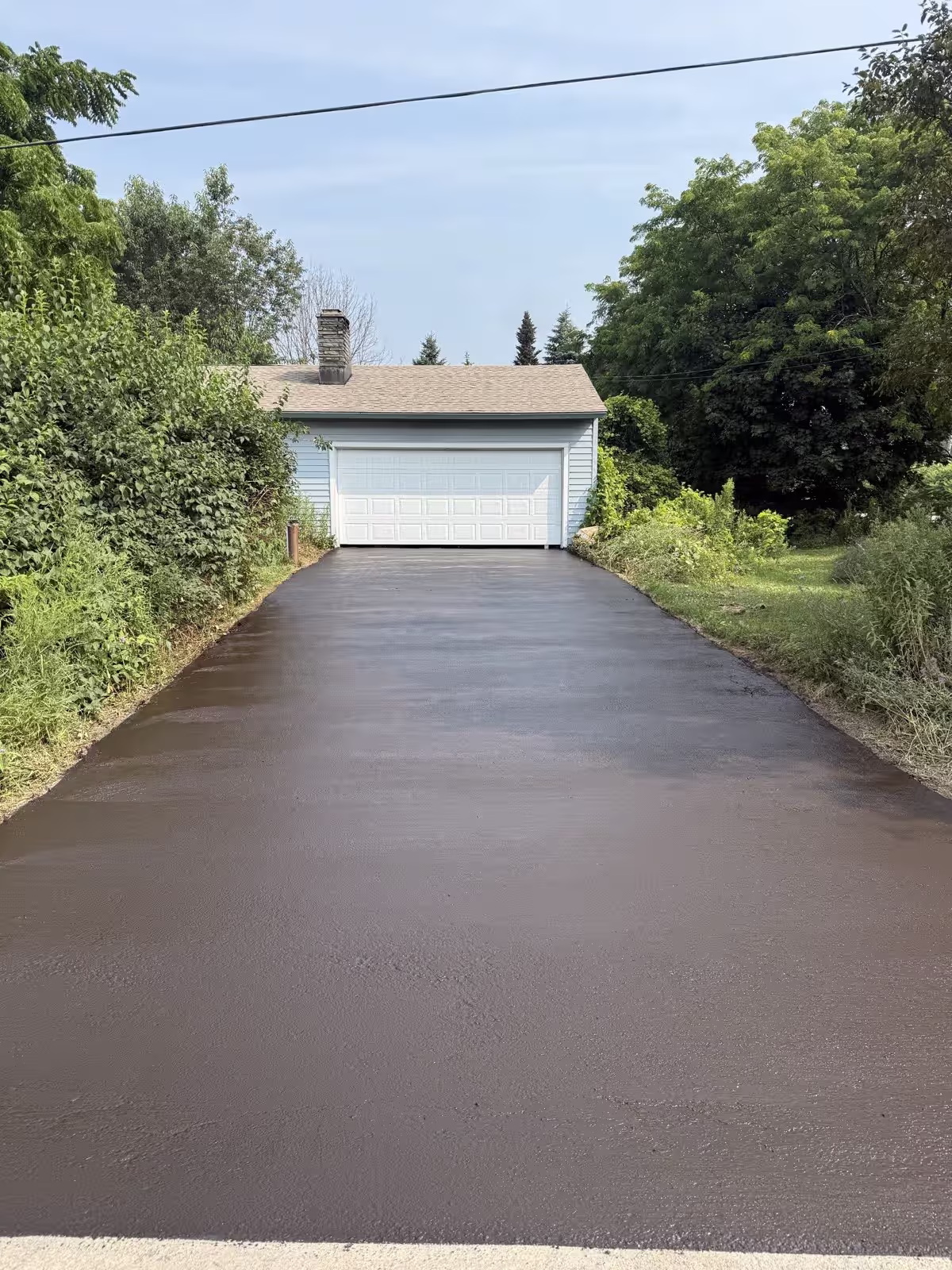A just-sealed driveway, the dark coat still wet and matte before it cures, leading to a white garage