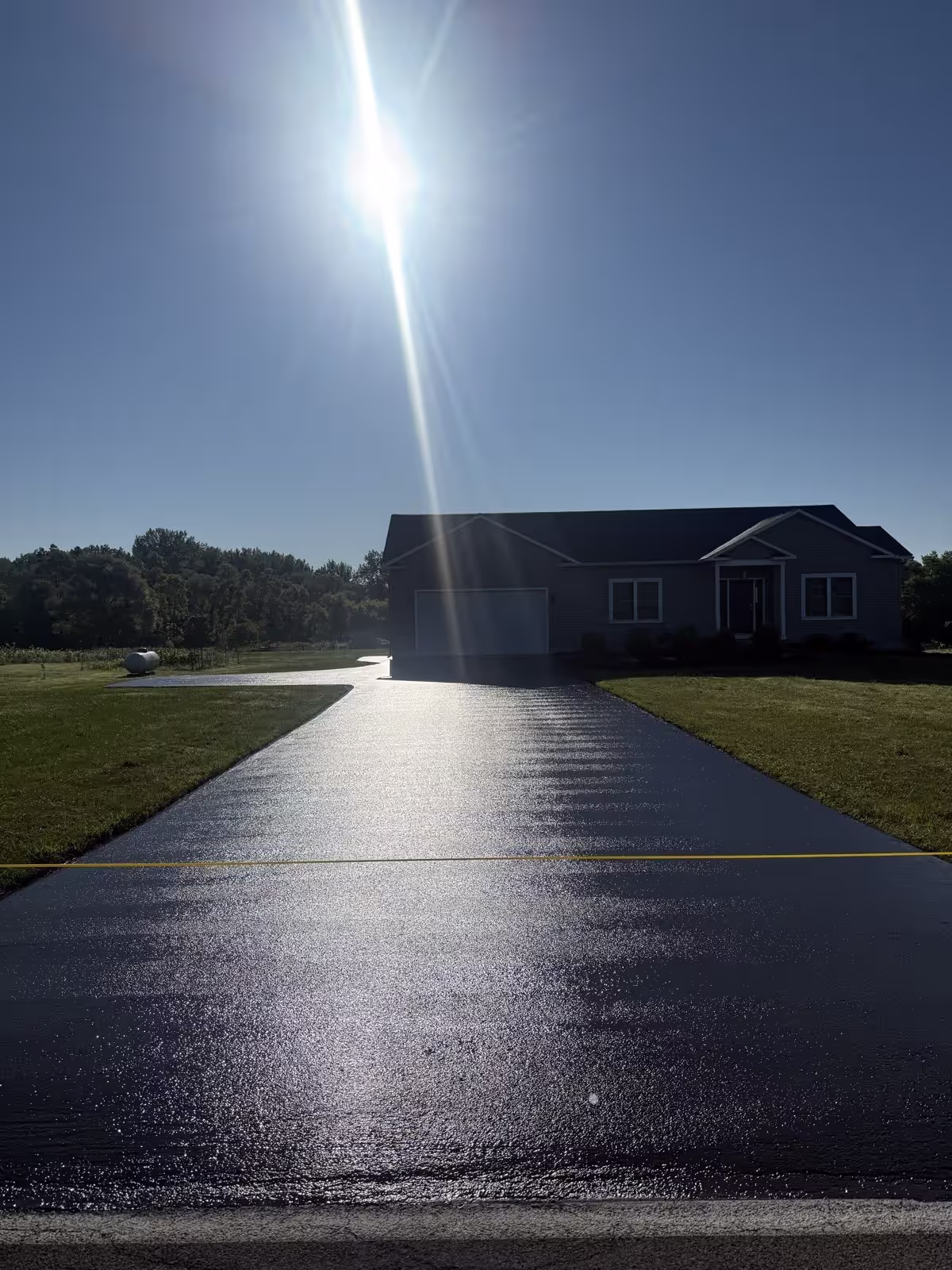 A black sealed driveway beside a gray two-tone house, lit by bright sun under a bold blue sky